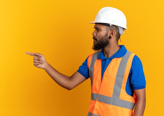 clueless young builder man in uniform with safety helmet looking and pointing at side isolated on orange wall with copy space