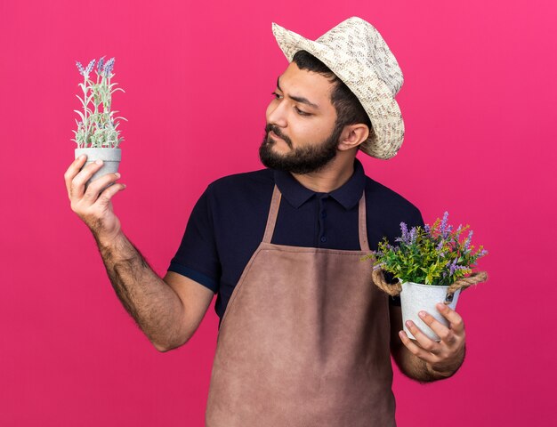 clueless young caucasian male gardener wearing gardening hat holding flowerpots isolated on pink wall with copy space