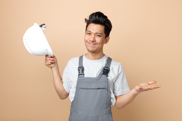 Clueless young construction worker wearing uniform holding safety helmet showing empty hand 