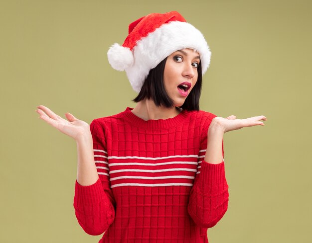 Clueless young girl wearing santa hat looking at camera showing empty hands isolated on olive green background