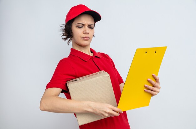 Clueless young pretty delivery woman holding cardboard box and looking at clipboard 