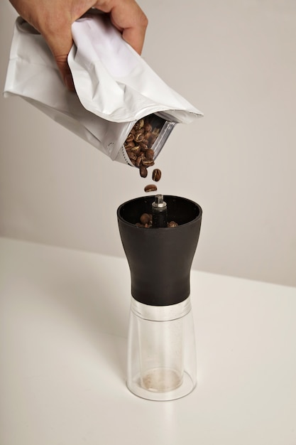 Free photo coffee beans fall into a compact slim manual grinder standing on white table from a white foiled bag