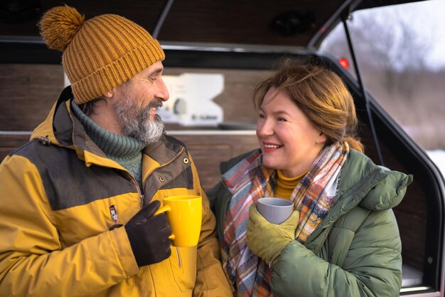 Coffee break during journey mature couple relax moment together at mini camper Middle aged couple drink from cups standing nest to a camper Family adventure trip fun time Family journey concept