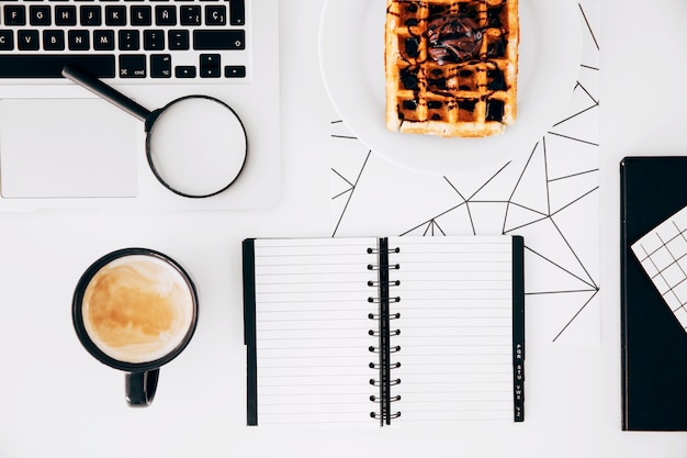 Coffee cup; laptop; magnifying glasses; spiral notepad and waffles with chocolate on plate against white desk