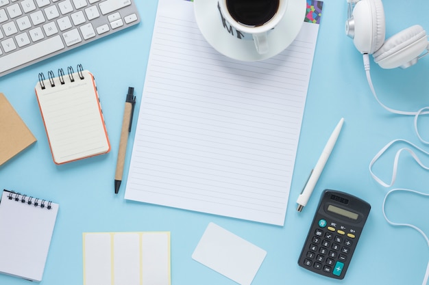 Coffee cup on single line page; spiral notepad; pen; keyboard; headphone against blue background