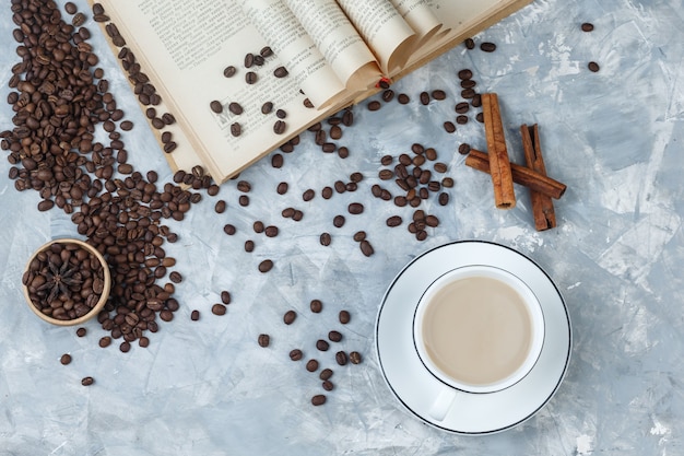 Coffee in a cup with coffee beans, book, cinnamon sticks top view on a grey plaster background