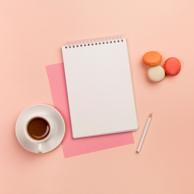 Coffee cup with spiral notepad,white pencil and macaroons on colored backdrop