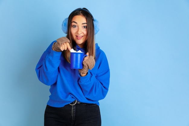 Coffee time. Caucasian woman's portrait on blue studio background. Beautiful female model in warm clothes. Concept of emotions, facial expression, sales, ad. Winter mood, Christmas time, holidays.