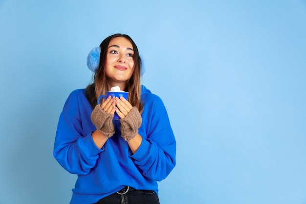 Free Photo coffee time. caucasian woman's portrait on blue studio background. beautiful female model in warm clothes. concept of emotions, facial expression, sales, ad. winter mood, christmas time, holidays.