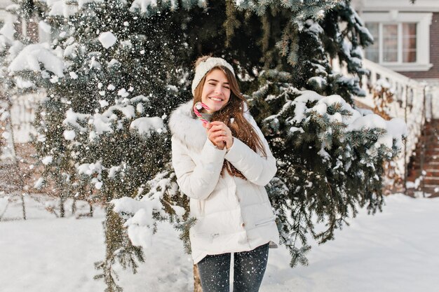 Cold winter weather in city and funny amazing enjoyed girl with lollypop on fri tree background. Warm clothes, happy mood, falling snow around, positive emotions, new year, sweet frozen winter time