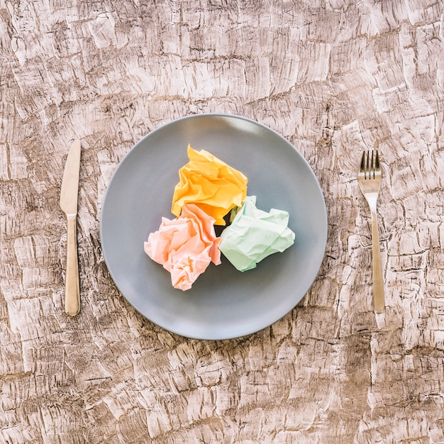 Free photo colorful crumpled papers on plate between fork and kitchen knife over wooden surface