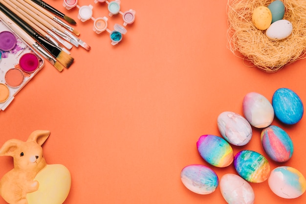 Free photo colorful easter eggs; nest; paint brushes; paint water color box and rabbit statue against orange backdrop