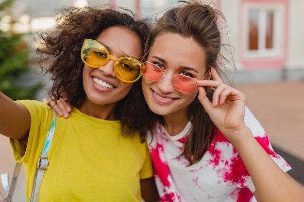 Colorful portrait of happy young girls friends smiling sitting in street taking selfie photo on mobile phone, women having fun together