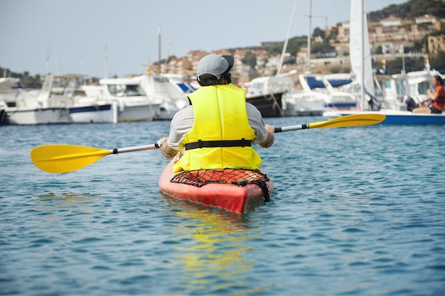 Free photo colorful shot of canoeing young man in a yellow swimming vest with paddle stopped in the air. good-looking landscape of lovely harbor with white yachts and nearby city houses on the mountain.