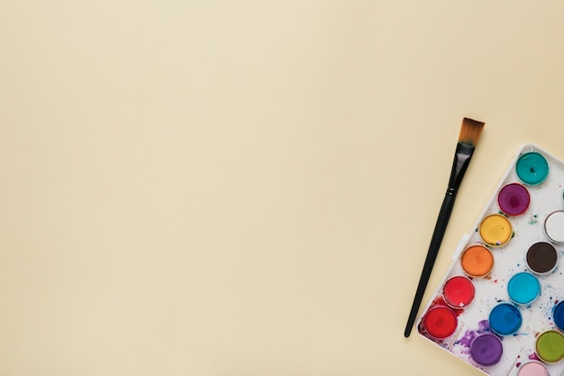 Colorful watercolor palette and paint brush on beige backdrop