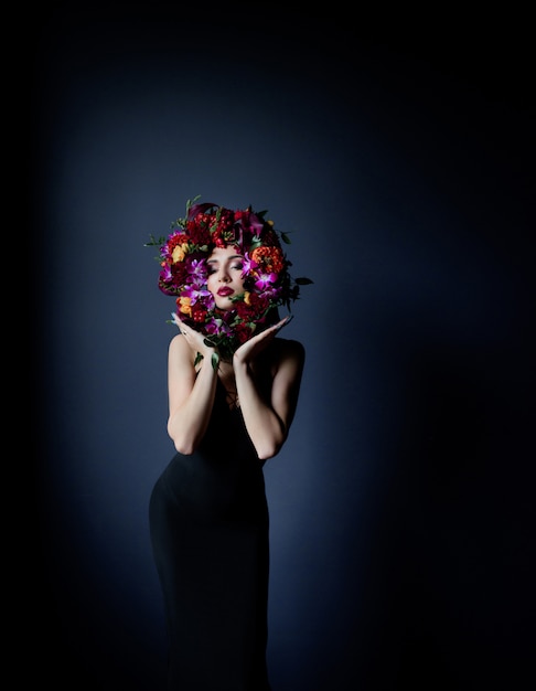 Colourful circle made of fresh flowers on the beautiful girl's face, woman dressed in black tight dress on the dark blue background