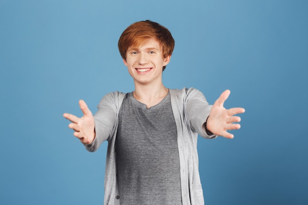 Come here, I'll give you a hug. Positive emotions. Redhead attractive joyful young guy in trendy grey clothes pulling hands with bright smile and happy expression.