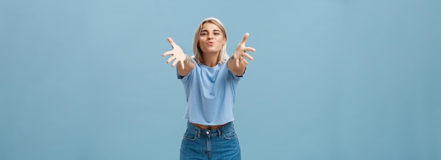 Free photo come to mama portrait of friendly joyful and enthusiastic caucasian female student in trendy outfit pulling hands towads camera and folding lips to hug and give passionate kiss smiling over blue wall