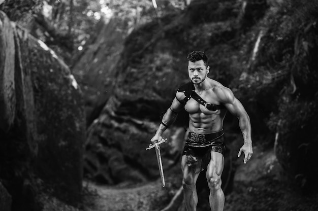 Free photo come at me. monochrome shot of a young manly gladiator holding a sword ready to fight near the rocks