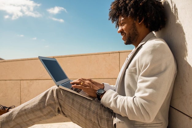 Comfortable African American man working in open air. Man in suit with beard using laptop. Sitting at terrace or rooftops. Working, manager, technology concept