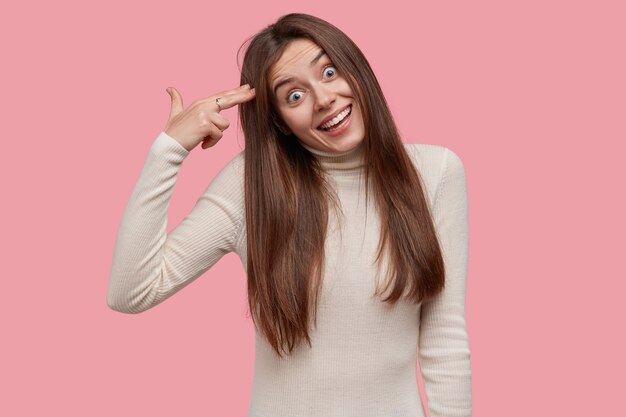 Comic beautiful young woman with joyful expression, keeps fingers on temples, pretends doing suicide gesture, dressed casually