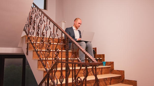 Company manager using laptop overworking on deadline sitting on stairs in finance building. Executive manager doing overtime at job on staircase businesspeople working in modern financial workplac e.