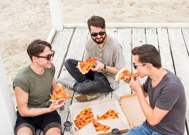 Company of young guys eating pizza on beach