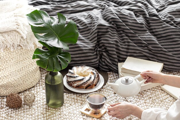 Composition of a cup of tea, homemade cupcake and decorative leaves in a vase against the background of a cozy bed.