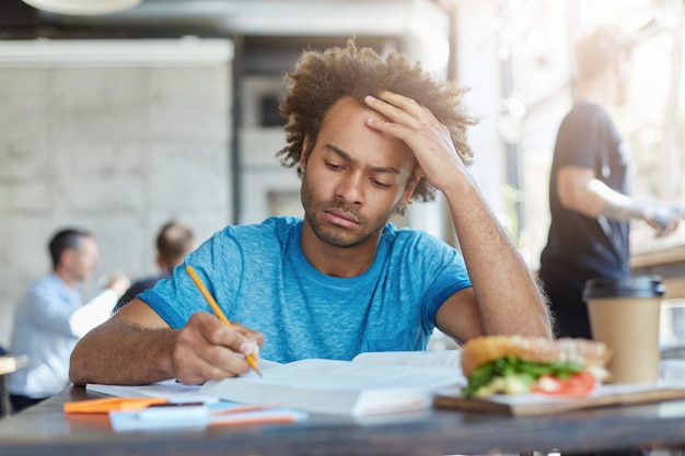 Concentrated casually dressed dark-skinned unshaven student studying at coffee shop, writing down in exercise book, making research or preparing for examination at college, having serious look