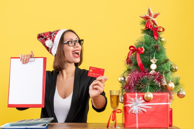 Concentrated charming lady in suit with santa claus hat and eyeglasses showing bank card and document in the office on yellow isolated 