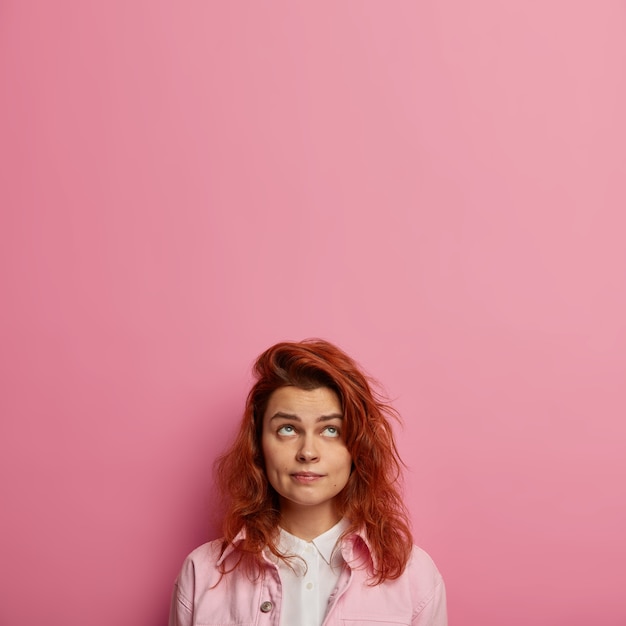 Free photo concentrated european woman with ginger hair, healthy skin, looks upwards, wears pink clothes