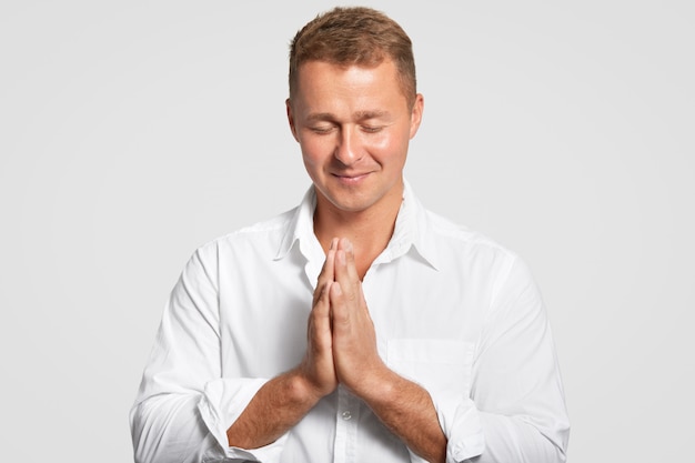 Free photo concentrated faithful young male keeps hands in praying gesture, closes eyes, begs for something desirable, hopes dreams come true, dressed in white shirt, stands alone