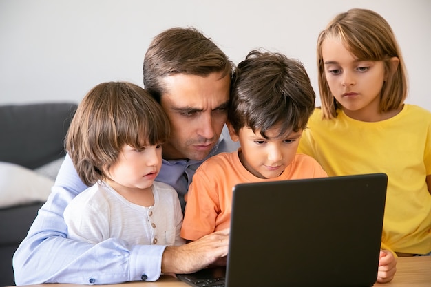 Free photo concentrated father and serious kids looking at screen. caucasian middle-aged dad typing on laptop screen and children watching his work. fatherhood, childhood and digital technology concept