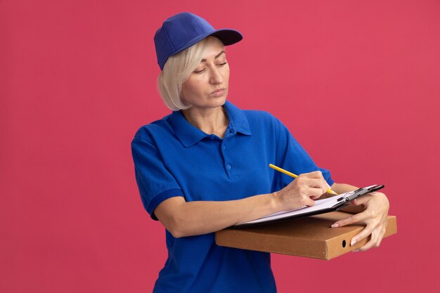 Concentrated middle-aged blonde delivery woman in blue uniform and cap writing with pencil on clipboard holding pizza package isolated on pink wall with copy space