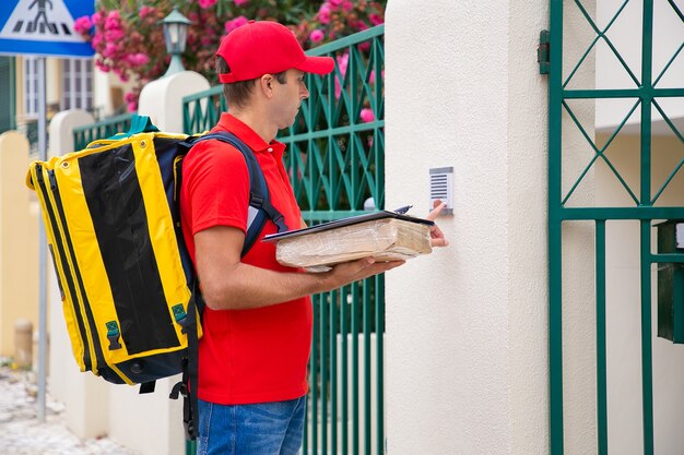 Concentrated postman ringing doorbell and holding parcel and clipboard. Handsome courier in red uniform carrying backpack, standing outdoors and delivering order. Delivery service and post concept