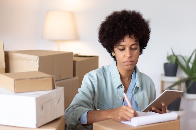 Concentrated woman making notes about package. Serious young logistics clerk using digital tablet while working at warehouse. Storeroom concept