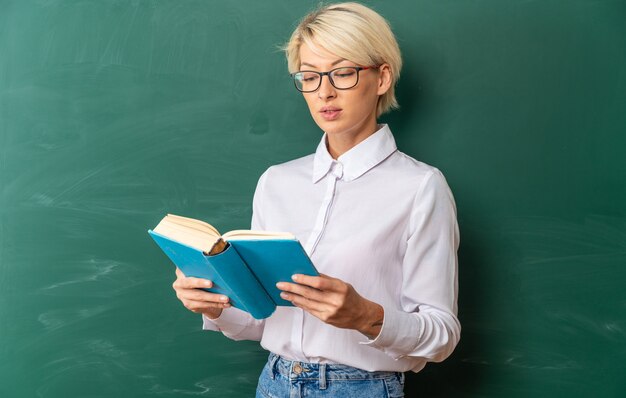 concentrated young blonde female teacher wearing glasses in classroom standing in front of chalkboard holding and reading book