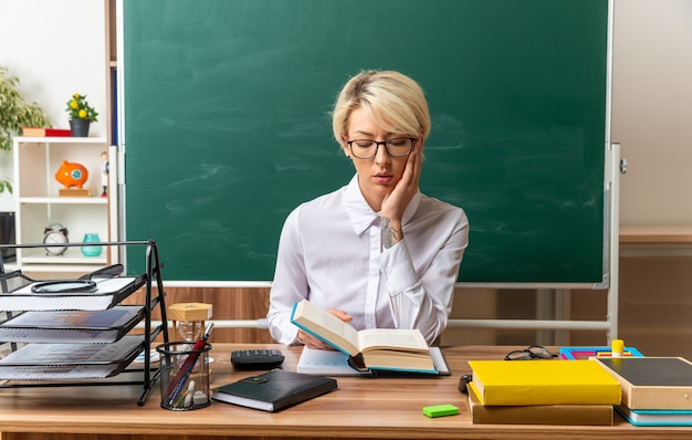 concentrated young blonde female teacher wearing glasses sitting at desk with school supplies in classroom reading book keeping hand on face