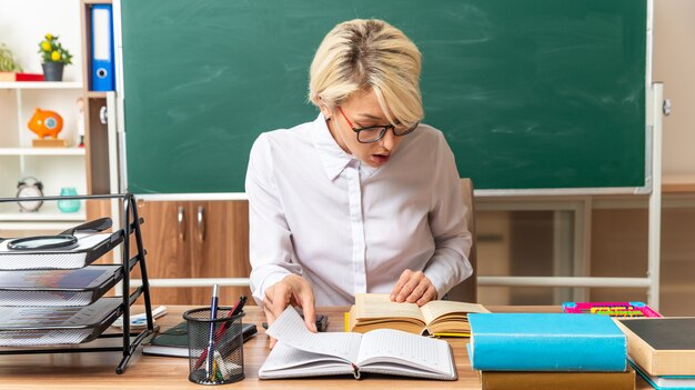 concentrated young blonde female teacher wearing glasses sitting at desk with school tools in classroom grabbing note pad keeping hand on open book looking at book