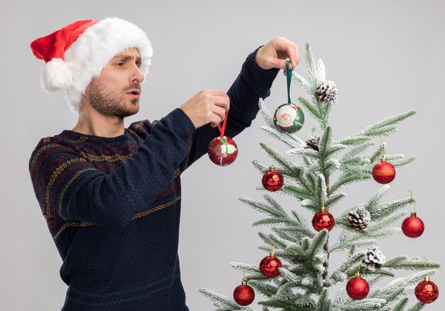 Free Photo concentrated young caucasian man wearing christmas hat standing near christmas tree decorating it with christmas ornament balls isolated on white wall