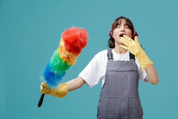 Concentrated young female cleaner wearing uniform bandana and rubber gloves holding feather duster keeping hand in front of mouth getting ready to sneeze with closed eyes isolated on blue background