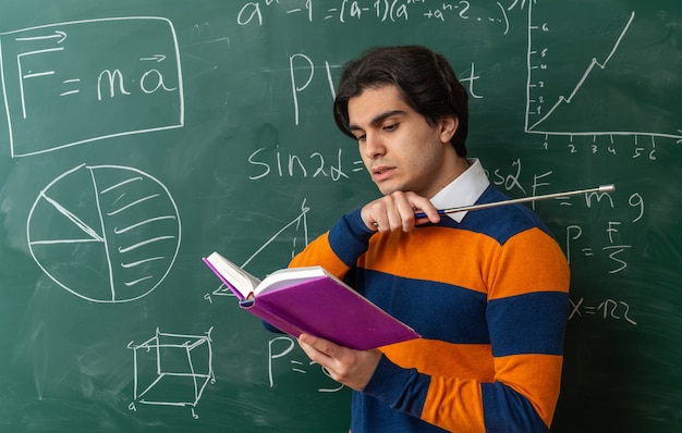 Free photo concentrated young geometry teacher standing in profile view in front of chalkboard in classroom reading book pointing at side