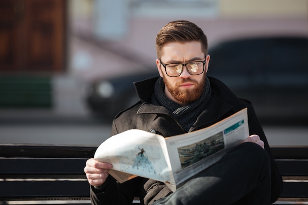 Free photo concentrated young man sitting on bench and reading newspaper outdoors