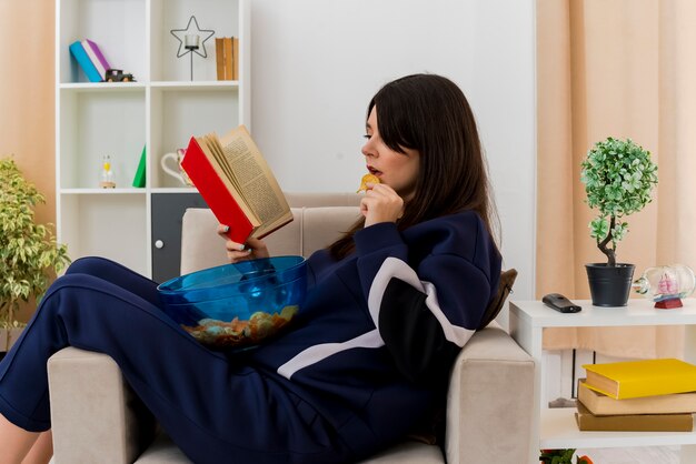 Concentrated young pretty caucasian woman sitting on armchair in designed living room holding bowl of chips on legs eating potato chip and reading book