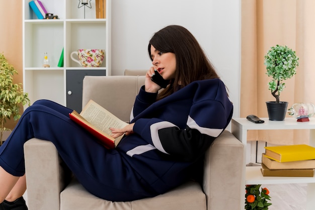 Concentrated young pretty caucasian woman sitting on armchair in designed living room talking on phone with book on legs looking at book and touching it