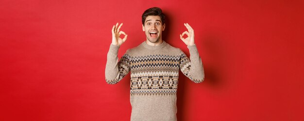 Concept of christmas celebration winter holidays and lifestyle Portrait of handsome man in sweater looking amazed and showing okay signs guarantee or recommend something good red background