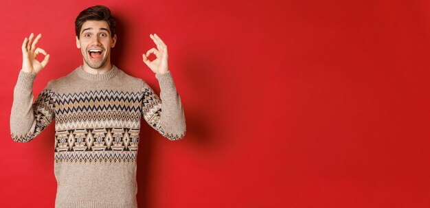 Concept of christmas celebration, winter holidays and lifestyle. Portrait of handsome man in sweater, looking amazed and showing okay signs, guarantee or recommend something good, red background.