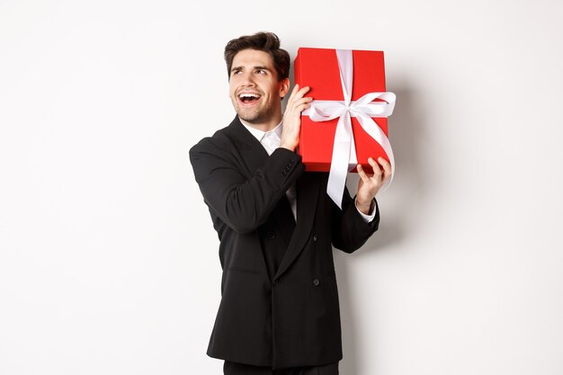 Concept of christmas holidays, celebration and lifestyle. Image of excited man enjoying new year, shaking gift box to guess what inside, standing against white background.