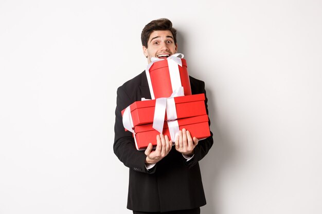 Concept of christmas holidays, celebration and lifestyle. Image of happy man in suit carry presents for new year, holding boxes with gifts and smiling, standing against white background