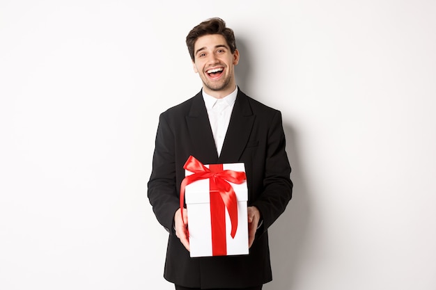 Free photo concept of christmas holidays, celebration and lifestyle. joyful handsome man in black suit, holding xmas gift and smiling, standing against white background.